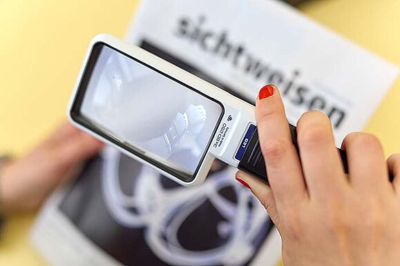 Nahaufnahme der Hände einer Person, die mithilfe einer Lupe ein Magazin liest. Sie hält die Lupe mit der rechten Hand, mit der linken das Magazin auf dem "sichtweisen" steht.
