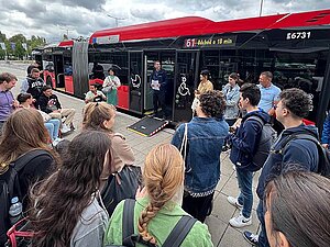 Eine Gruppe junger Menschen steht an einer Bushaltestelle in Bratislava und hört einem Mann zu, der vor einem roten Linienbus mit geöffneter Tür spricht. Eine Rampe für barrierefreien Zugang ist ausgeklappt. Der Bus zeigt die Liniennummer 61 und die Abfahrt in 18 Minuten an.