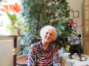 Veronika K. kann keine Gesichter mehr erkennen Veronika K. sitzt vor ihren Pflanzen und neben einer Katzenstatue und lächelt.