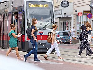 Blindenleitsysteme sind wichtige Hilfsmittel im Straßenverkehr. © Ludwig Schedl Personen kreuzen an Zebrastreifen die Straße, auch eine junge Frau mit Langstock, sunkler Brille und Rucksack. Dahinter wartet eine Straßenbahn mit der Aufschrift: "5, Westbahnhof"