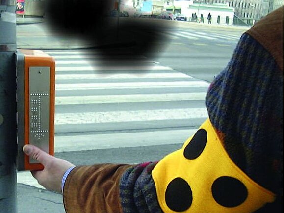 Ein Mensch mit Blindenabzeichen steht an einer Ampel. Über dem Zebrastreifen sieht man dunkle Flecken, wie es bei der Augenerkrankung AMD, Altersbedingter Makula-Degeneration der Fall ist.