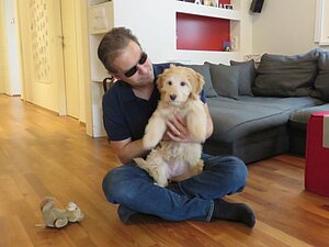 Ein Mann sitzt im Schneidersitz am Boden. Er hält einen braunen Hund mit beiden Händen.