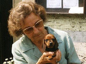 Helene Lantzberg Eine ältere Dame mit großer Sonnenbrille hält einen braunen Dackelwelpen in den Armen
