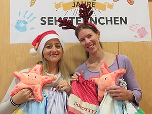 Gemeinsam haben wir es geschafft! Zwei Mitarbeiterinnen der Hilfsgemeinschaft zeigen Kuschelseesterne und Turnbeutel mit Namen drauf her. Dabei Tragen sie Weihnachtsmützen.