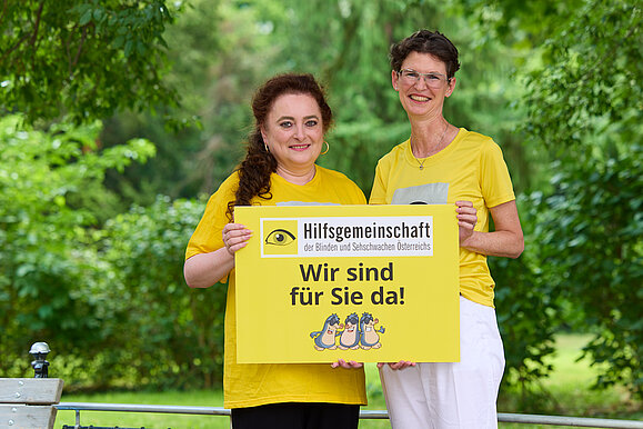 Zwei Frauen in gelben Shirts stehen vor Büschen, halten ein Schild und lächeln in die Kamera