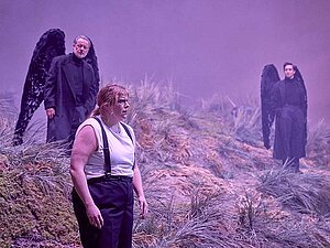 Theaterszene mit drei Personen auf einer nebligen, hügeligen Bühne mit Gras und Moos. Im Vordergrund steht eine Person mit weißem Hemd, schwarzen Hosenträgern und nachdenklichem Ausdruck. Dahinter befinden sich zwei Personen in schwarzen Gewändern mit großen schwarzen Engelsflügeln – eine links und eine rechts – in ruhiger, ernster Haltung.