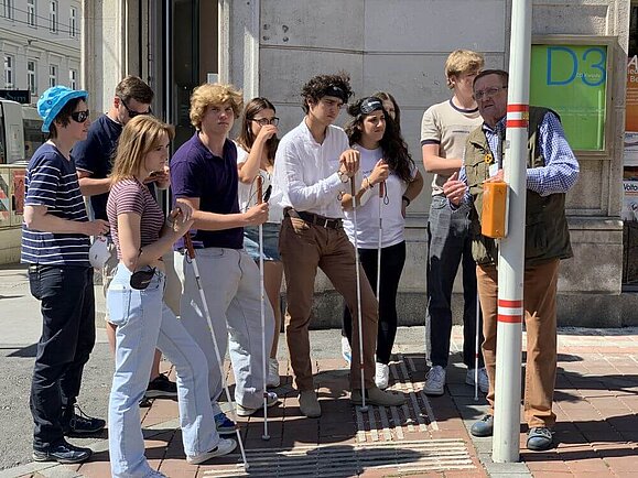 reiter_Schulungen_Tourismus_Schulklasse Mann mit Blinden-Kennzeichnung erklärt mehreren Jugendlichen mit Augenbinde und Langstock die Funktionen der Akustikampel