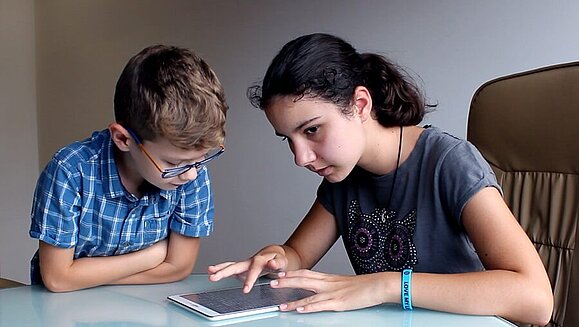 Maks spielt mit seiner blinden Schwester Mia „Schiffe versenken“, Copyright: Feelif Maks sitzt mit seiner blinden Schwester Mia vor dem Tablet, Mia führt ihren Zeigenfinger über das Tablet , Copyright: Feelif