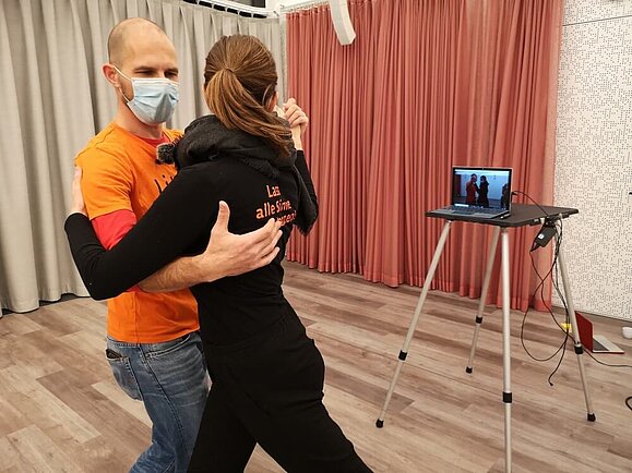 Lucia und Jörg beim Unterrichten eines Tango-Argentino-Online-Kurses. Die beiden stehen in Paartanzhaltung vor einem Laptop und erklären den tanzbegeisterten zu Hause eine Schrittsequenz. Copyright: Lillis Ballroom Zwei Personen stehen in Paartanzhaltung vor einem aufgeklappten Laptop der die beiden filmt.