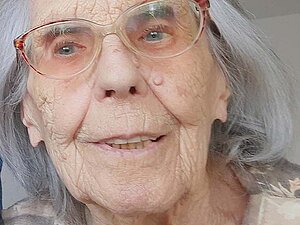 Hildegard Graf Portrait einer älteren Dame mit silbergrauen, schulterlangen Haaren. Sie trägt eine Brille mit rotem Rahmen und ein T-Shirt mit einem Blumenmuster in dezenten Farben.