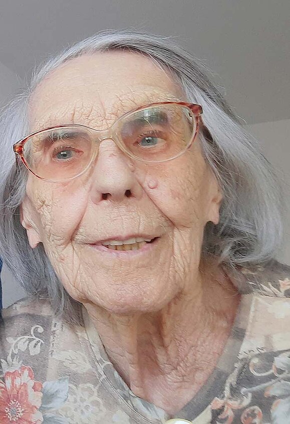 Hildegard Graf Portrait einer älteren Dame mit silbergrauen, schulterlangen Haaren. Sie trägt eine Brille mit rotem Rahmen und ein T-Shirt mit einem Blumenmuster in dezenten Farben.