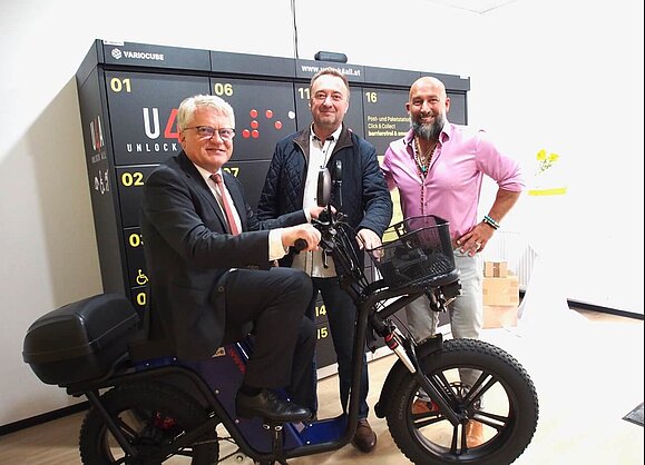 Das neue E-Lastenbike wird ebso mit vorgestellt Drei Herren mit schwarzem E-Lastenbike vor Paketbox