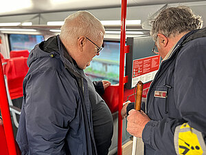 Zwei Männer stehen in einem modernen Zug vor einem Knopf. Beide tragen Jacken, einer hält einen Taststock und trägt eine gelbe Armbinde. Er drückt den Knopf, während ihn beide betrachten. 