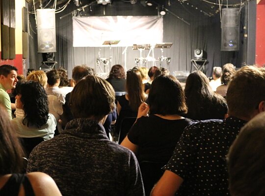 AUDIAMO Live-Hörspiel Mehrere Menschen stehen mit dem Rücken zur Kamera vor einer Bühne.