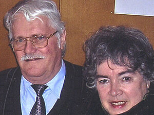 Monika und Herbert Neumann Ein älteres Ehepaar steht lächelnd, eng nebeneinander und blickt in die Kamera.