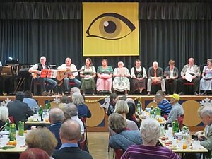 Auf der Bühne ist die Singgruppe Kunterbunt. Die Mitglieder sitzen an mehreren Tischen und folgen dem Programm.