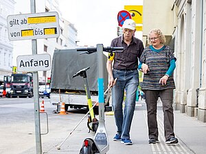älterer Mann mit Langstock eingehakt bei einer Frau geht auf E-Scooter mitten am Gehsteig zu