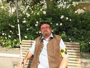 Fritz K. ist hochgradig sehbehindert und engagiert sich für die Hilfsgemeinschaft. Fritz K. sitzt auf einer Bank im Park, in der Hand einen Langstock, am anderen Arm eine Armbinde.