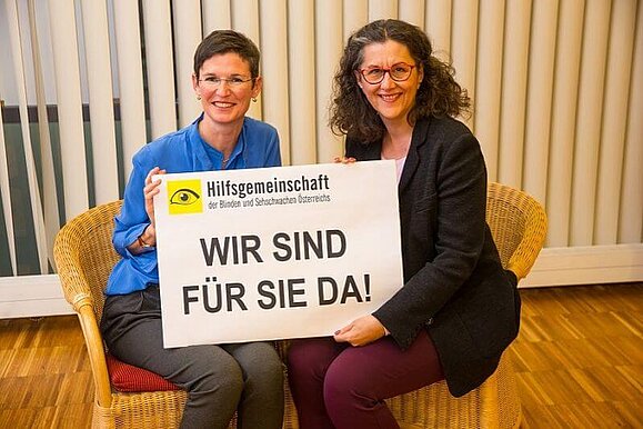 Wir sind für Sie da!, Copyright: Franz Pfluegl Zwei Mitarbeiterinnen der Hilfsgemeinschaft sitzen auf je einem Stuhl und halten gemeinsam lächelnd ein Schild mit den Worten "Wir sind für Sie da!" in die Kamera.