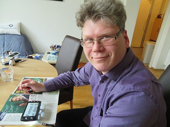 Peter G. sitzt an einem Tisch mit einer elektronischen Lupe. Peter G. sitzt an einem Tisch mit einer elektronischen Lupe.