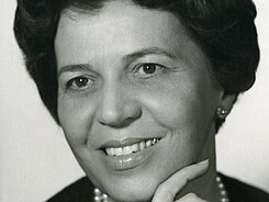 Augustine Mayer Schwarz-weiß Foto einer lächelnden Dame mittleren Alters, die ihre Hand ans Kinn hält und nicht direkt in die Kamera blickt