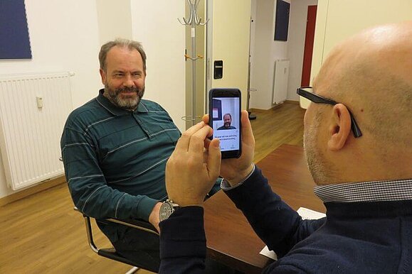 Die App beschreibt den fotografierten Mann so: „53 year old man with Gray hair and a beard looking happy.“ Mitarbeiter Daniele Marano fotografiert mit seinem Smartphone einen Mann mit Bart.“