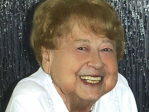 Waltraude Köckeis kam durch ihre eigene Sehbehinderung mit der Hilfsgemeinschaft in Kontakt. Foto: privat. ältere Frau mit weißem Shirt und dunkelblonden Haaren lächelt in die Kamera, Hintergrund in silber