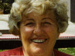 Wilhelmine Frischauf Eine ältere Dame mit Dauerwelle hält ein Glas in der Hand und lächelt fröhlich in die Kamera, hinter ihr sieht man einen Teil eines alten Bauernhauses