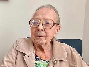 Ältere Frau mit Brille und beiger Jacke sitzt auf Stuhl