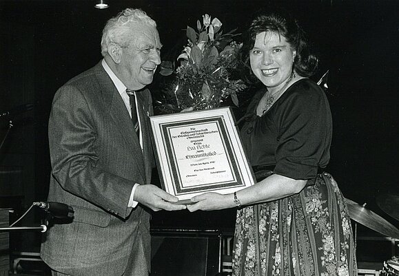 Prof. Robert Vogel überreicht Eva Fichte die Urkunde zur Ehrenmitgliedschaft. schwarz-weiß Foto: älterer Mann in Anzug überreicht Frau mit Föhnfrisur und Blumenrock Urkunde und Blumenstrauß