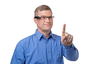 Mann in blauem Hemd hält seinen Zeigefinger in die Luft, auf seinem Kopf eine Brille mit OrCam, Copyright: OrCam VIDEBIS