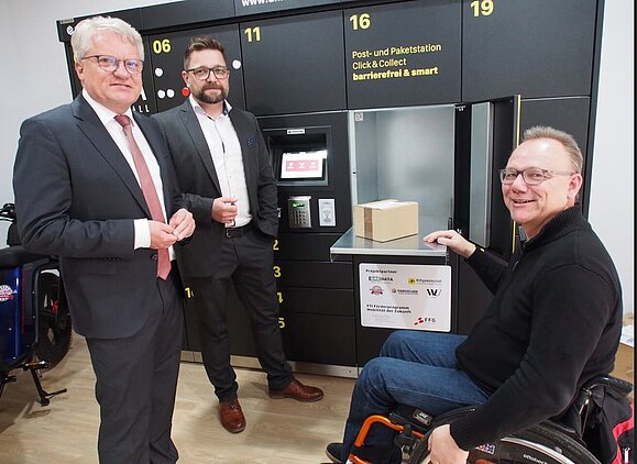 Die innovative Paketbox wurde bereits genauer unter die Lupe genommen Rollstuhlfahrer mit zwei elegant gekleideten Herren vor Paketbox
