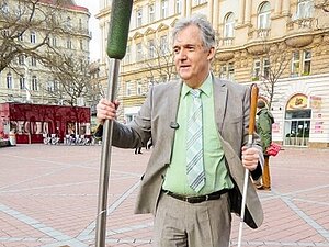 Ein Mann steht auf einem großen Platz, hält in der linken Hand einen Blindenstock und ertastet mit der rechten Hand eine Stange.