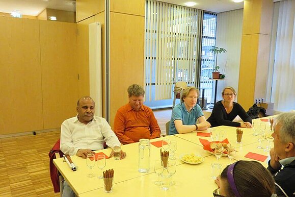 Gespannt lauschen die Teilnehmenden den Worten der Fachfrau. 4 Männer und eine Frau sitzen an einem gedeckten, langen Tisch mit Snacks und Gläsern.