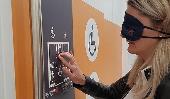 Taktile Übersichtspläne geben sehbeeinträchtigten Menschen Orientierung und ermöglichen so mehr Selbstständigkeit. Eine Frau mit Augenbinde ertastet einen taktilen weißen Orientierungsplan, der mit dunkler Farbe hinterlegt ist.