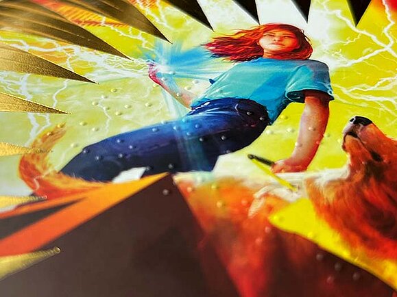 Nahaufnahme des Buchcovers „Ella Jones vs the Sun Stealer“. Zu sehen ist ein Mädchen mit rotem Haar in dynamischer Pose neben einem Hund, umgeben von leuchtenden Blitzen und goldenen Strahlen. Auf dem Cover sind erhabene Punkte in Braille-Schrift zu erkennen, die fühlbar sind.