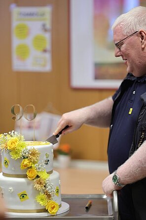 Ein Mann (Elmar Fürst) schneidet eine Torte an, auf welcher die Zahl 90 ist.
