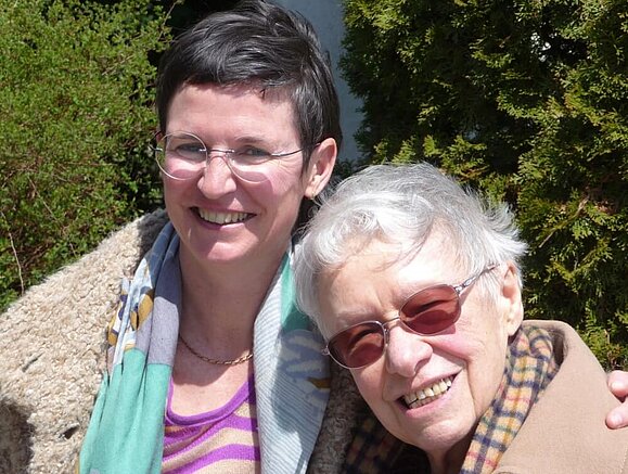 Zwei Frauen stehen nebeneinander, die ältere lehnt sich leicht an die jüngere, beide lächeln herzlich in die Kamera.