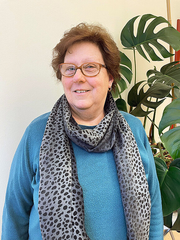 Frau lächelt in die Kamera, hat kurze braune Haare und eine Brille und trägt ein gemustertes graues Halstuch zu einem blauen Pullover.