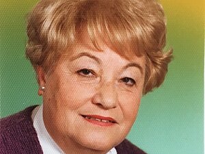 Josefine Prammer Eine Dame mit blonder Dauerwelle sitzt vor einem bunten Fotohintergrund und lächelt in die Kamera