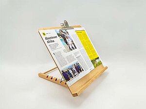 Lesepult aus Holz vor einem weißen Hintergrund. Das Pult ist eine Holzplatte mit Sockel unten, dahinter ein Ständer, der es erlaubt, den Winkel der Neigung einzustellen. Oben ist eine Klammer aus Metall befestigt, mit der ein Magazin, das aufgeschlagen auf dem Pult liegt, befestigt worden ist. 