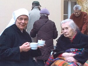Schwester Consulate mit Frau Fiala. Nonne mit Teetasse in der Hand und ältere Dame mit Decke schauen in die Kamera