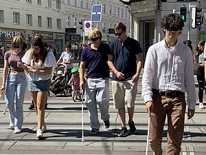 mehrere Junge Menschen, teils eingehakt mit weißem Stock und Augenbinde gehen über Zebrastreifen