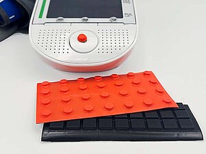 Klebepunkt auf einem Blutdruckmessgerät. Jeweils ein Bogen mit erhabenen orangen und schwarzen Klebepunkten, dahinter ein Gerät, auf dessen Bedienfeld ein orangener Punkt klebt.