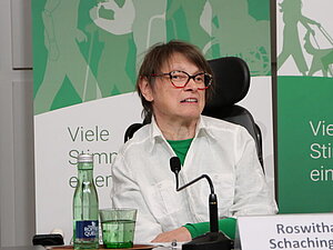Eine Frau mit braunem kurzen Haar und weißer Jacke sitzt auf einem Podium mit grünem Banner im Hintergrund.