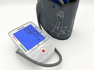 Nlutdruckmessgerät in silber mit blau leuchtendem Bildschirm und Knöpfen sowie anhängender grauer Manschette.