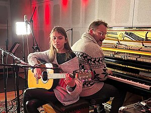 Eine junge Frau die Gitarre spielt sitzt Rücken an Rücken mit einem Mann, der eine elektronischen Orgel spielt, in einem Tonstudio. 