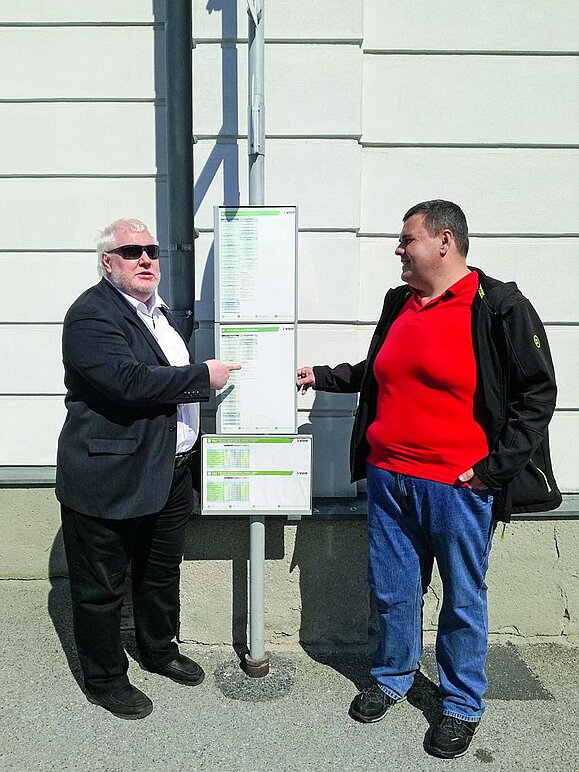 Elmar_Andreas_beim Lokalaugenschein Zwei Männer mit schwarzen Jacken zeigen auf ausgehängten Busplan