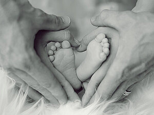 Vier Hände bilden ein Herz um die Füße eines Babys.
