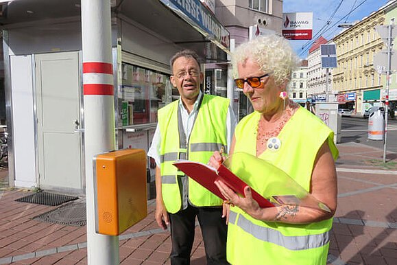 Ein Mann und eine Frau mit gelben Warnwesten stehen nebeneinander vor einem Masten mit einer akustischen Ampelanlage. Die Frau notiert etwas in einem Schnellhefter.